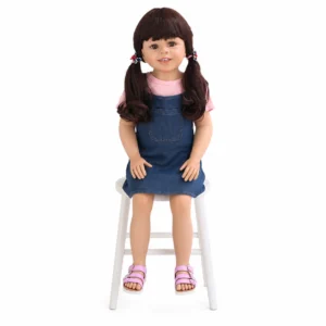 Lebensechte Puppe mit braunem Haar zu Zöpfen geflochten, trägt ein pinkfarbenes Hemd und ein Jeanskleid, sitzt auf einem weißen Stuhl.